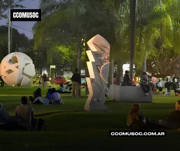 Parque nocturno con personas sentadas en el césped y esculturas iluminadas entre palmeras