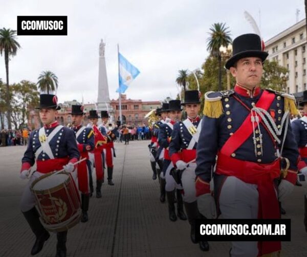 Desfile militar con granaderos de uniforme azul y rojo en una plaza, con un monumento al fondo